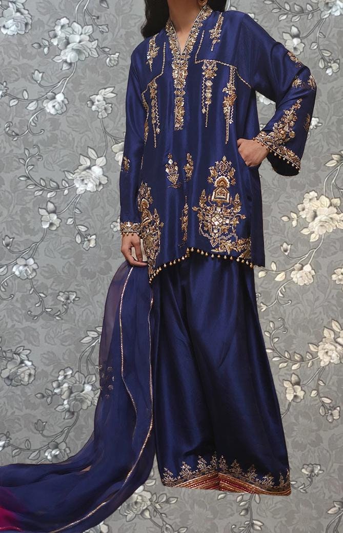 A woman wearing a blue and gold dress with intricate embroidery and patterns, standing against a gray floral background.