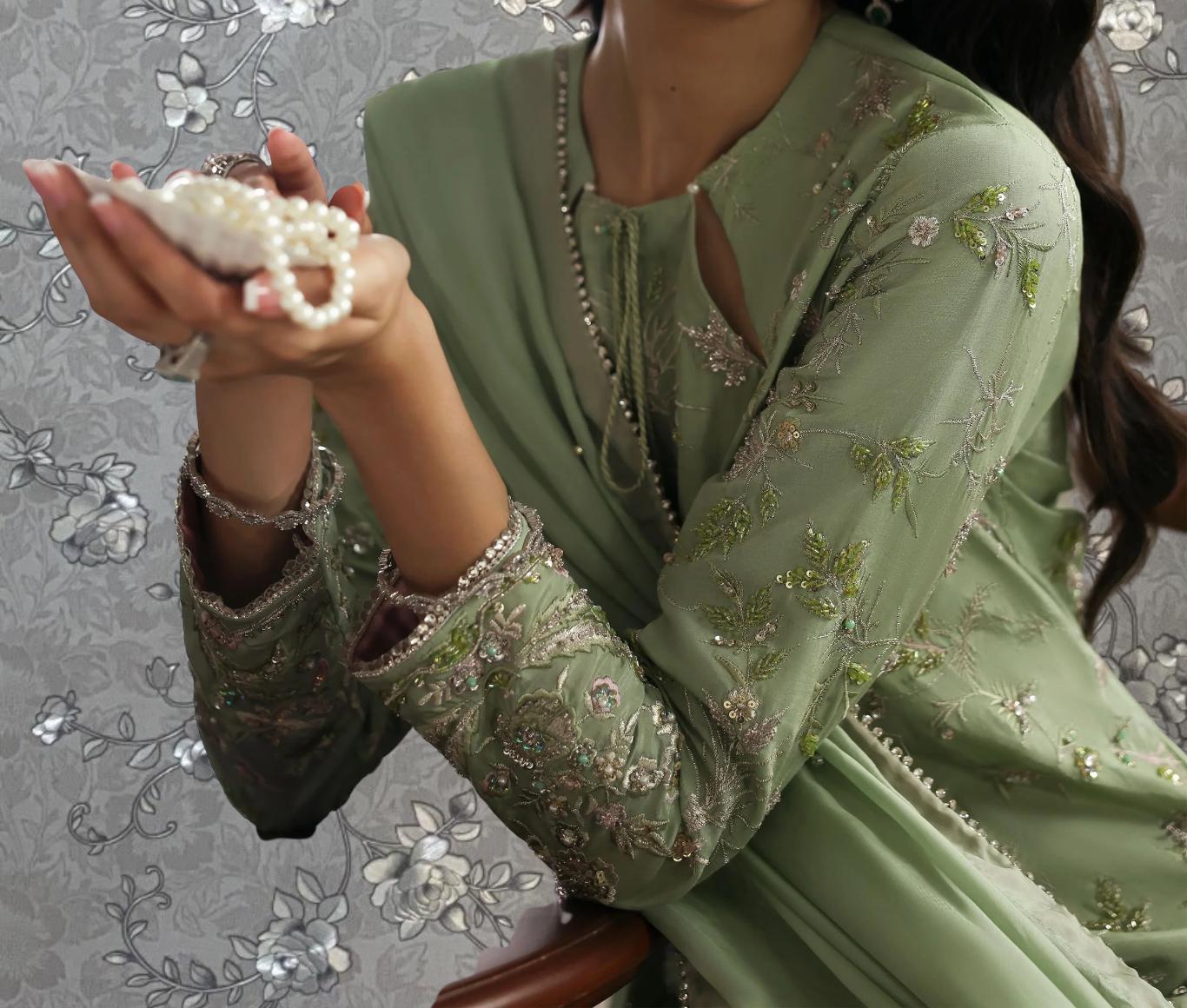 A woman in a green dress with intricate embroidery holds a small white object, possibly a pearl, in her hands.
