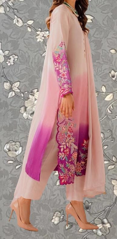 A woman wearing a long, flowing pink dress with intricate floral embroidery stands against a gray floral background.