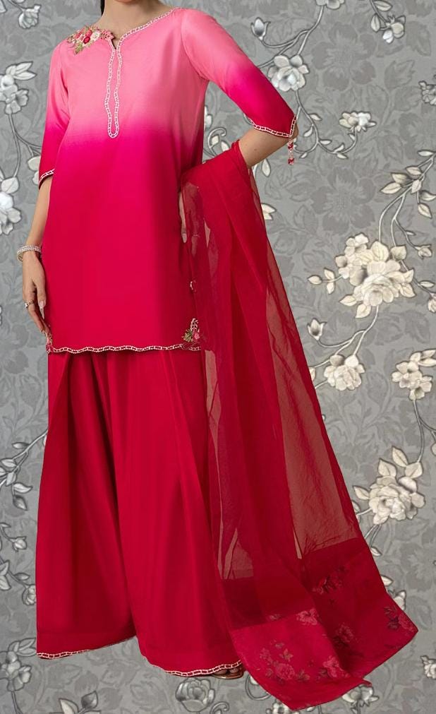 A woman wearing a pink and red dress with a matching red shawl, standing against a gray floral background.