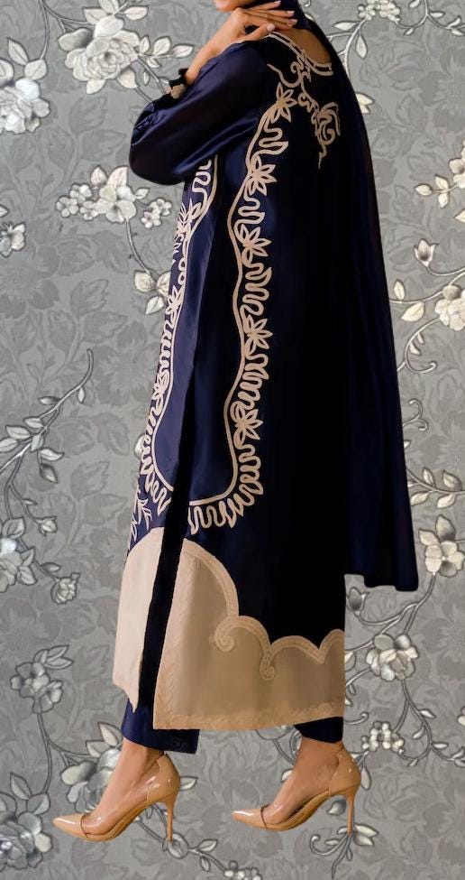 A person wearing a long navy blue dress with intricate white embroidery stands against a gray floral background.