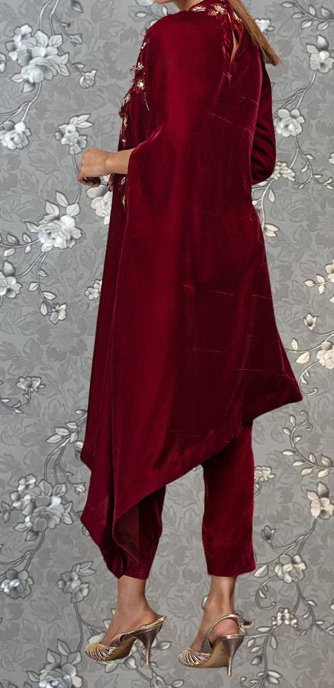 A person wearing a long, burgundy velvet dress with a high collar and a cape-like garment over the shoulders, standing against a gray floral background.