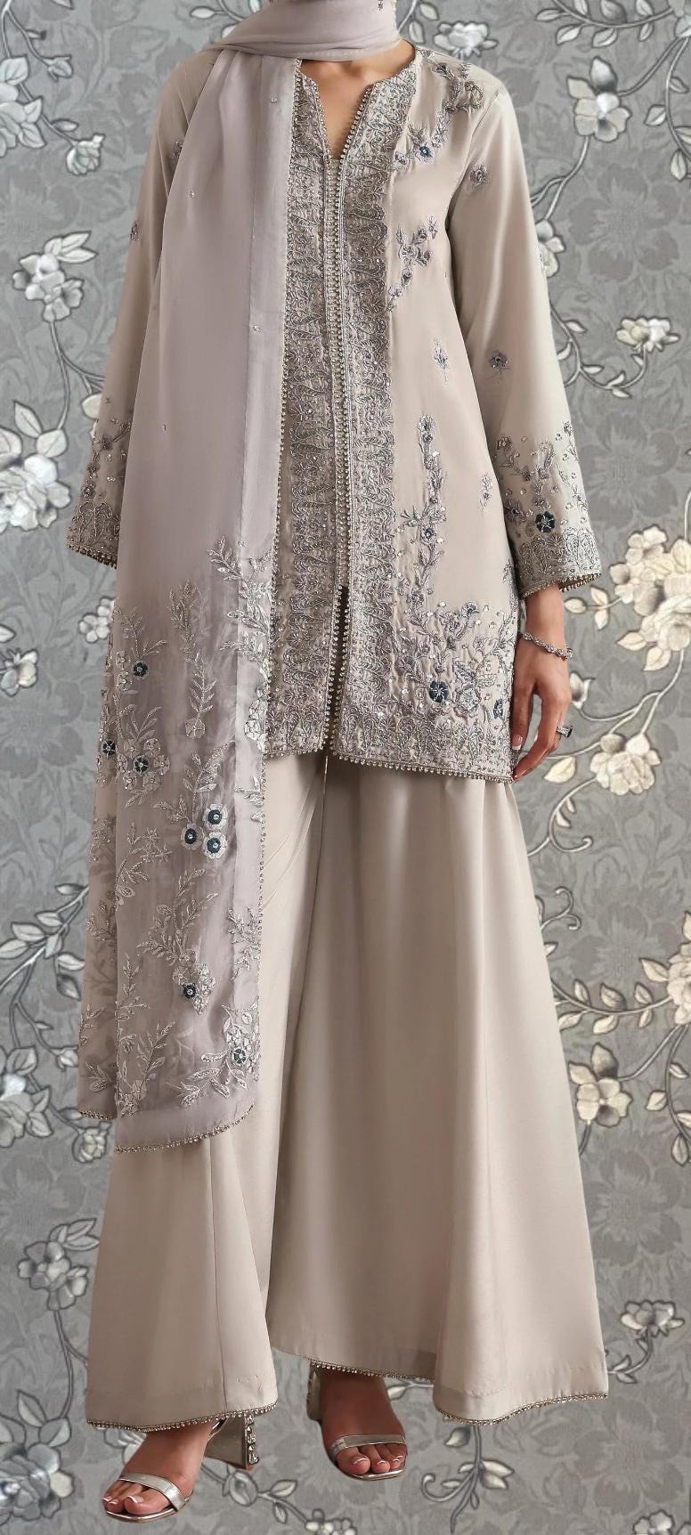 A woman wearing a beige dress with intricate embroidery and a matching beige scarf, standing against a gray floral background.