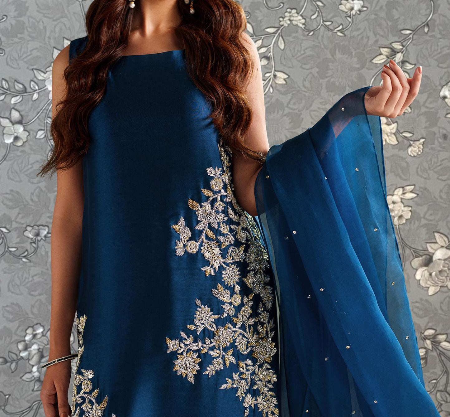 A woman wearing a blue dress with intricate embroidered designs, posing with her hand outstretched.