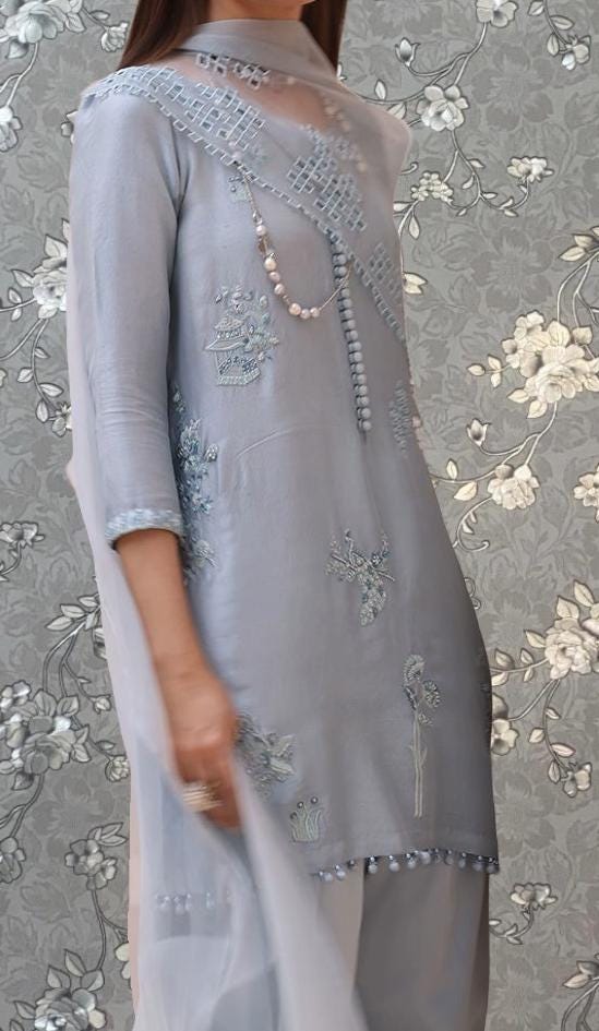 A woman wearing a gray dress with intricate embroidery and floral designs, standing against a floral patterned background.