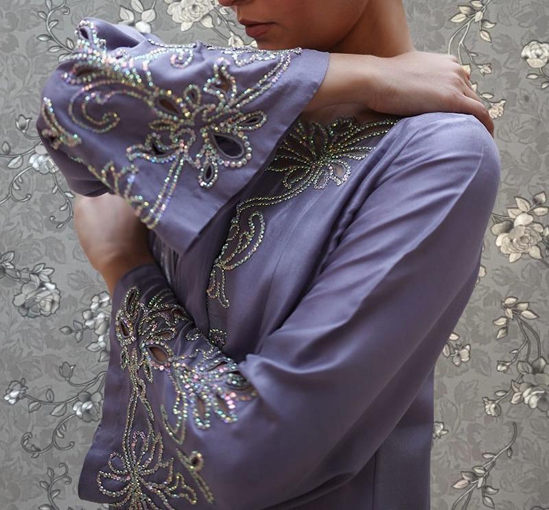 A person wearing a purple dress with intricate embroidered designs on the sleeves and collar stands against a floral patterned background.
