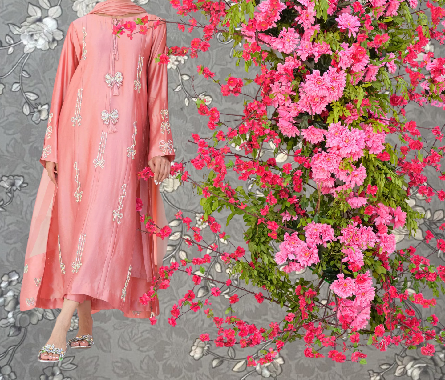 A woman wearing a pink dress with white floral embroidery stands next to a large floral arrangement of pink flowers.