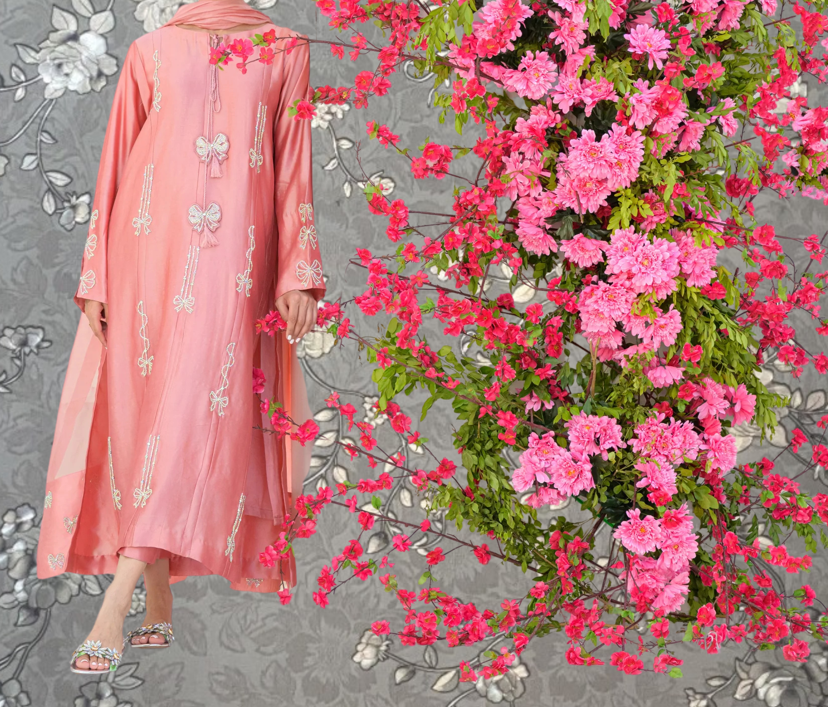 A woman wearing a pink dress with white floral embroidery stands next to a large floral arrangement of pink flowers.