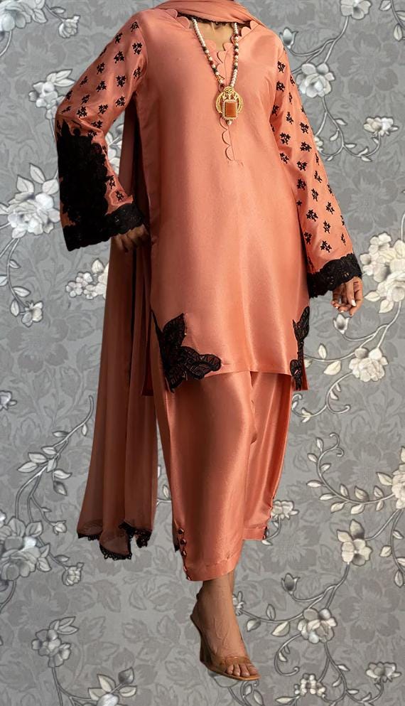 A woman wearing a peach-colored dress with black lace accents and a matching skirt, standing against a gray floral background.