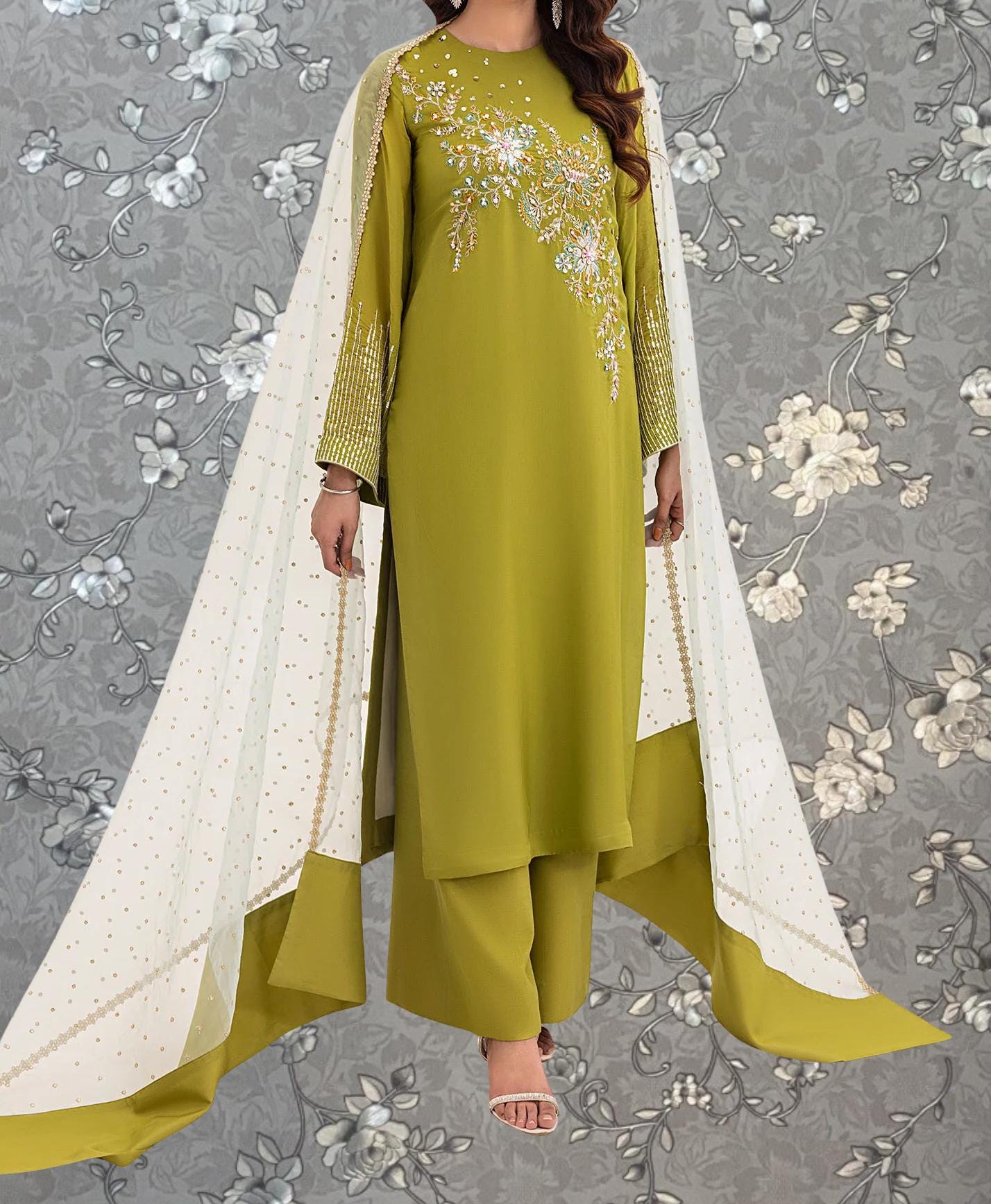 A woman wearing a green dress with gold embroidery and a white shawl, standing against a gray floral background.