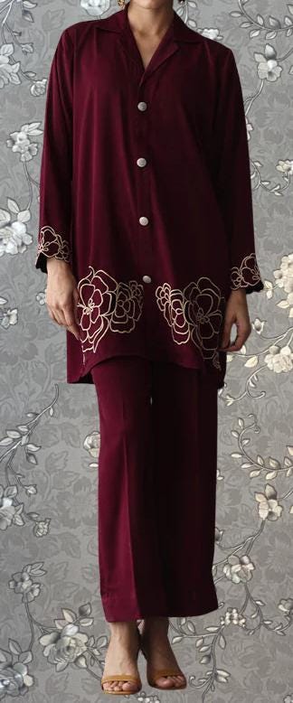 A person wearing a burgundy outfit with gold floral embroidery on the sleeves and pants, standing against a gray floral background.