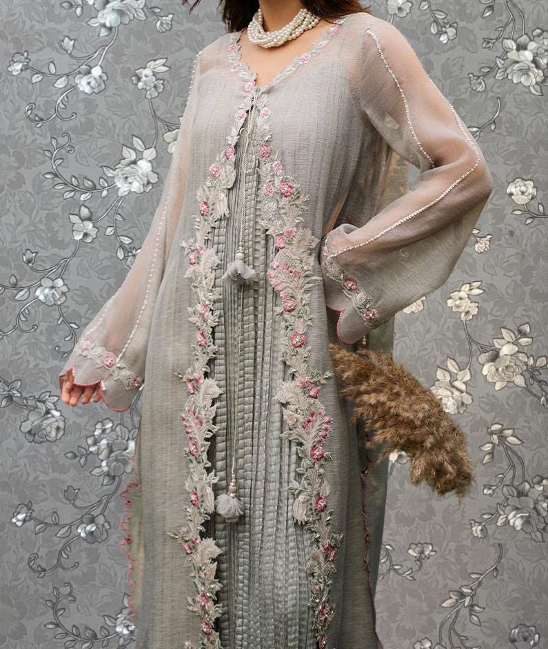A woman wearing a long, gray dress with intricate embroidery and floral patterns, posing against a gray floral background.