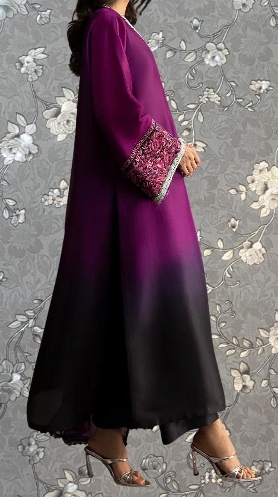 A woman wearing a long purple dress with a floral pattern on the sleeves and a matching purple and black skirt, standing against a gray floral background.