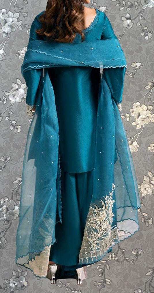 A woman wearing a teal-colored dress with a matching shawl, standing against a gray floral background.