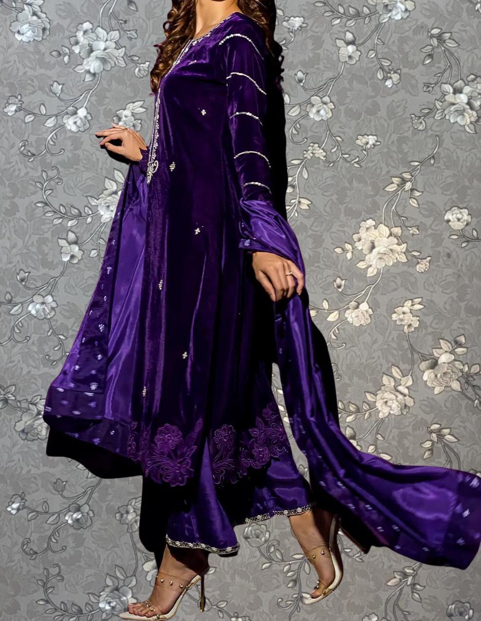 A woman wearing a long purple dress with intricate embroidery and a matching purple shawl is standing against a gray floral background.