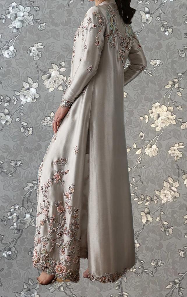 A woman wearing a long, elegant dress with intricate floral embroidery stands against a gray floral background.