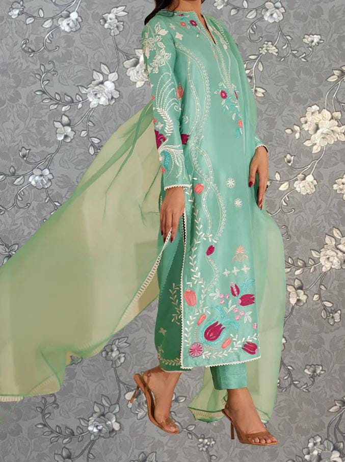 A woman wearing a long, mint green dress with intricate floral embroidery stands against a gray floral background.