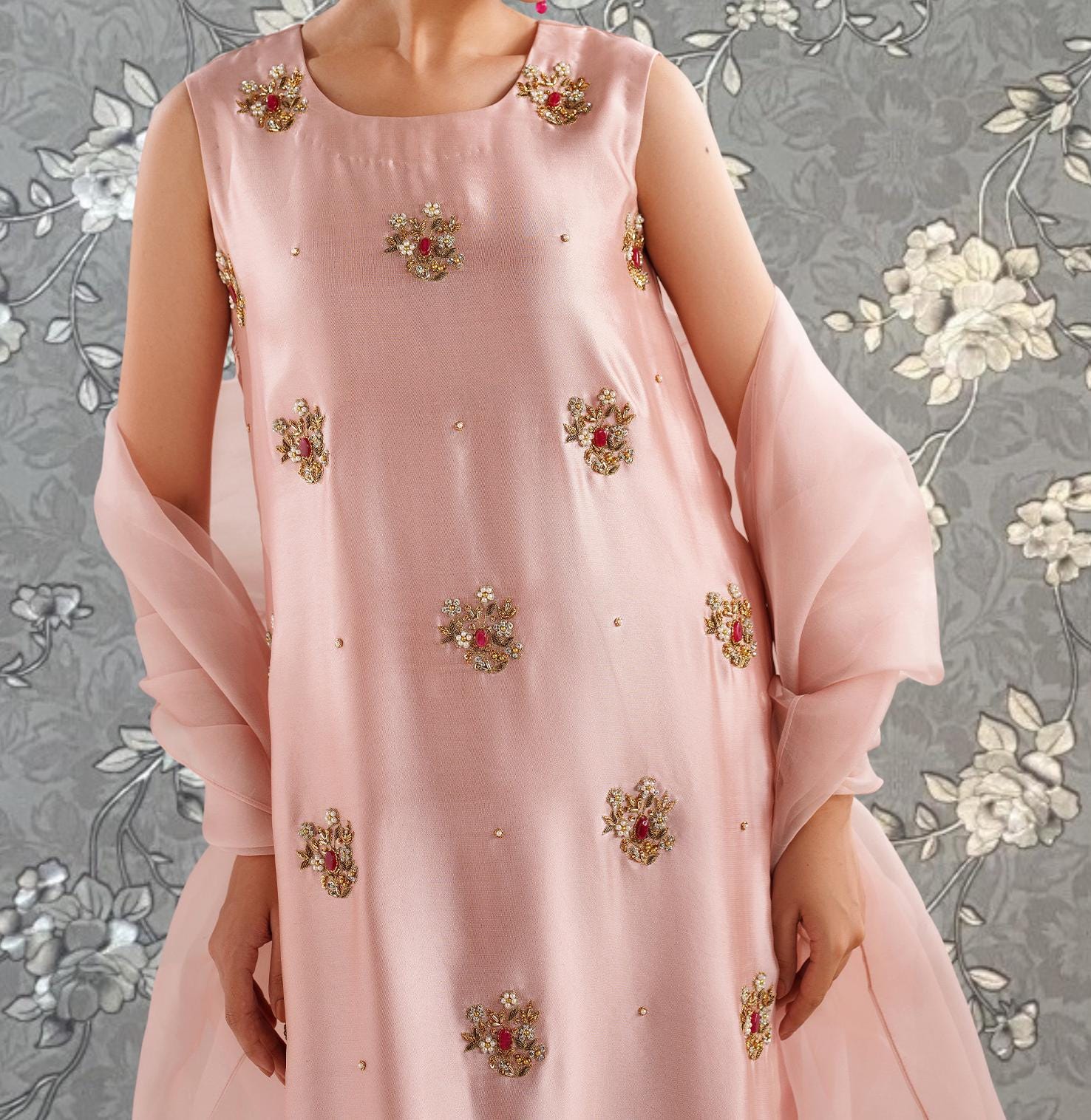 A woman wearing a pink dress with gold floral embroidery stands against a gray floral background.