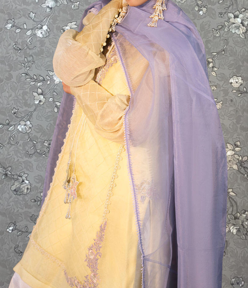 A person wearing a yellow and purple dress with a sheer purple shawl, standing against a gray floral background.