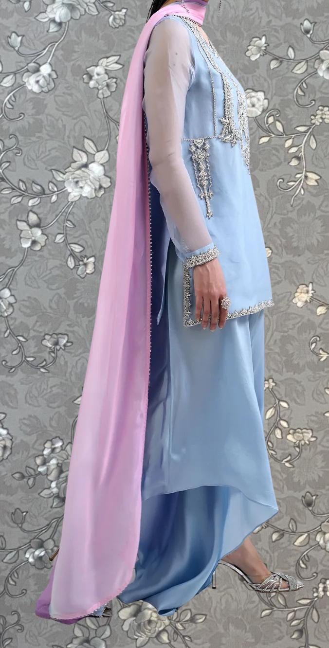 A woman wearing a light blue dress with a pink shawl, standing against a gray floral background.