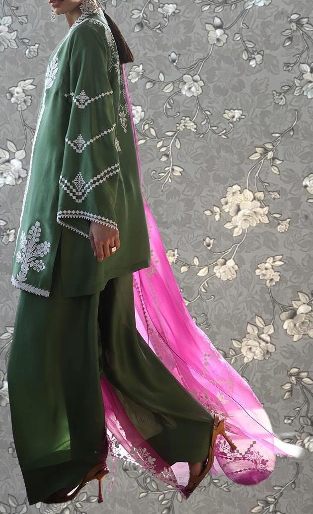 A person wearing a green and pink patterned outfit with a long pink shawl, standing against a gray floral background.