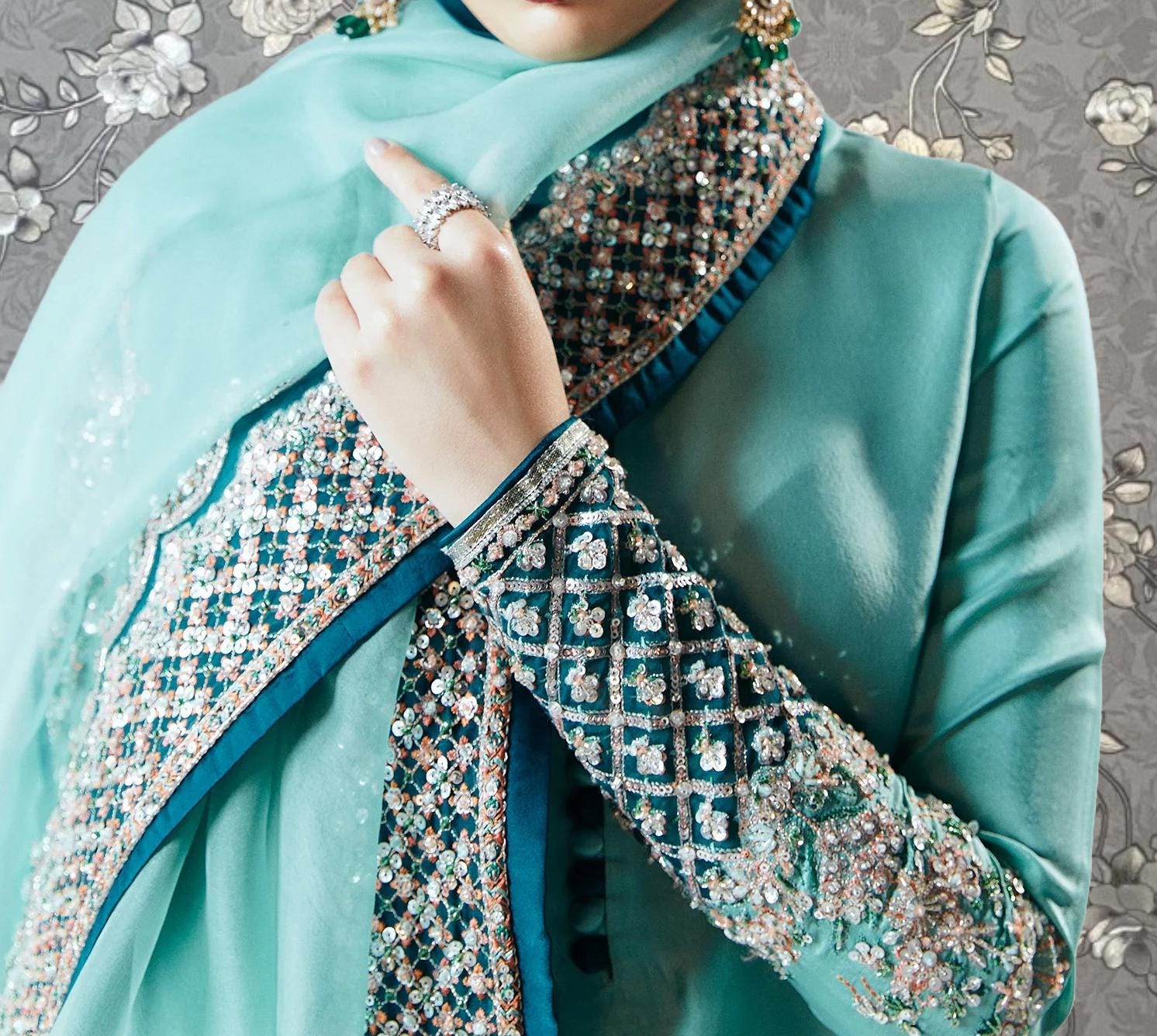 A close-up of a person wearing a turquoise dress with intricate embroidery and jewelry, including a ring on their finger.