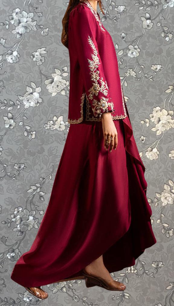 A woman wearing a long, red dress with intricate embroidery stands against a gray floral background.