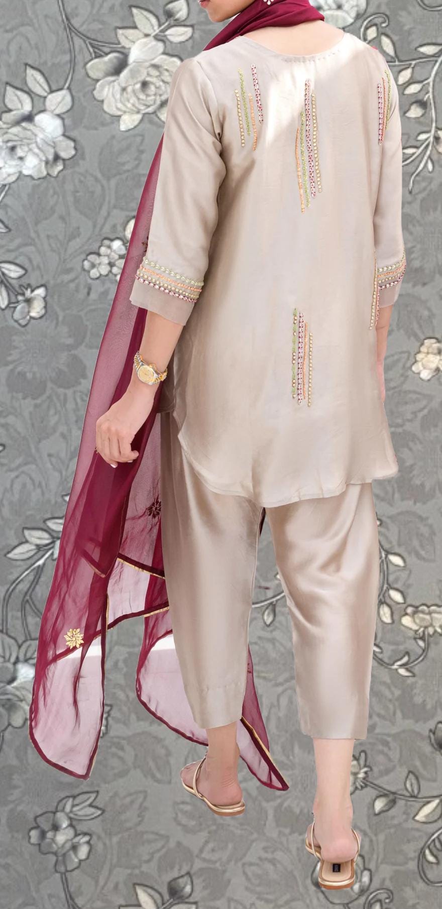 A person wearing a beige outfit with a red scarf is standing against a gray floral background.