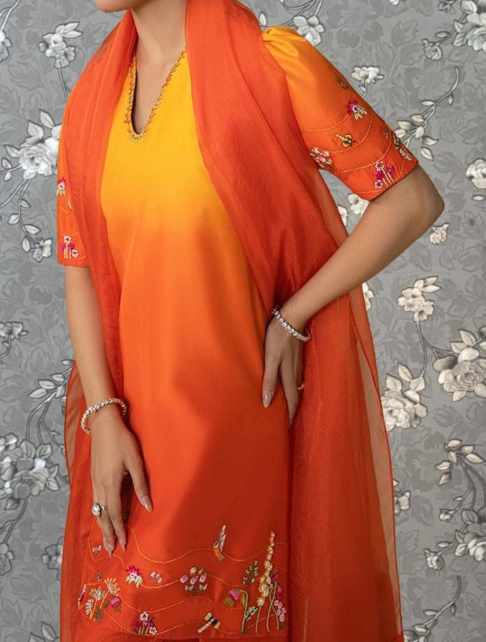 A woman wearing a vibrant orange dress with floral embroidery, posing against a gray floral background.