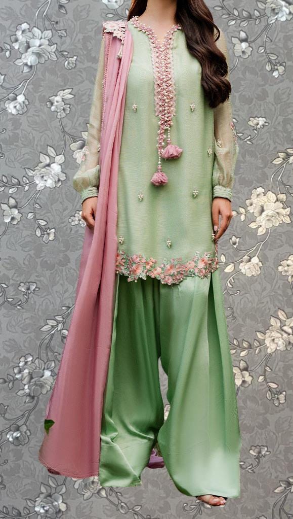 A woman wearing a green and pink dress with floral embroidery and a pink shawl, standing against a gray floral background.