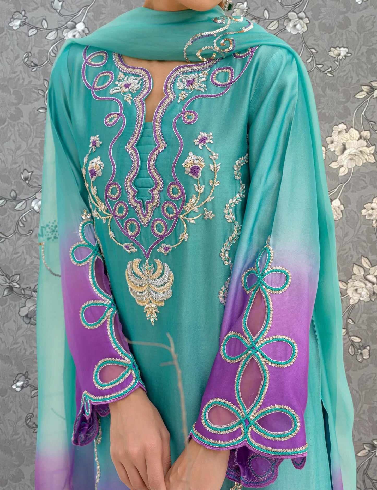A person wearing a turquoise and purple dress with intricate embroidered patterns on the sleeves and collar.