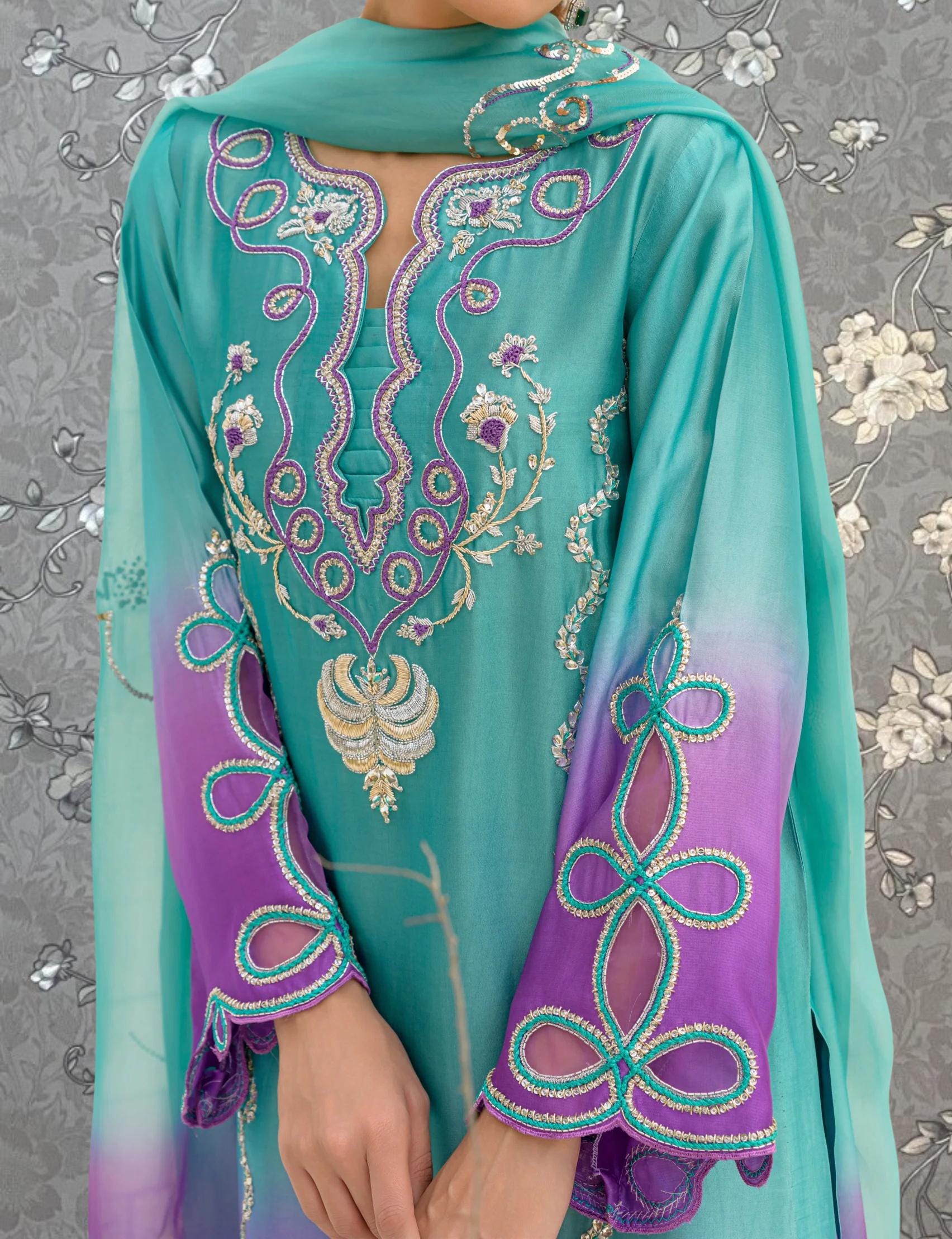A person wearing a turquoise and purple dress with intricate embroidered patterns on the sleeves and collar.