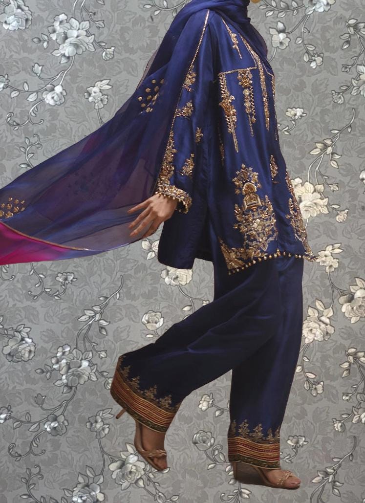 A person wearing a blue and gold outfit with intricate embroidery, standing against a gray floral background.