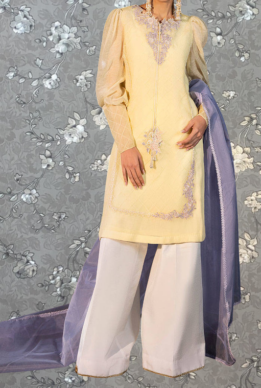 A woman wearing a yellow dress with a floral pattern and white pants stands against a gray floral background.