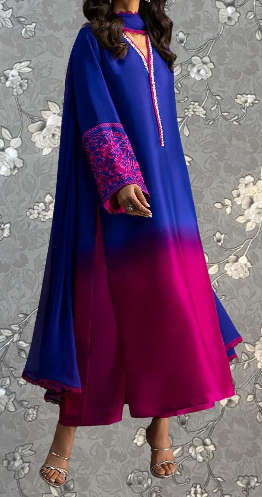 A woman wearing a long, flowing dress with a mix of blue and pink colors, standing against a gray floral background.