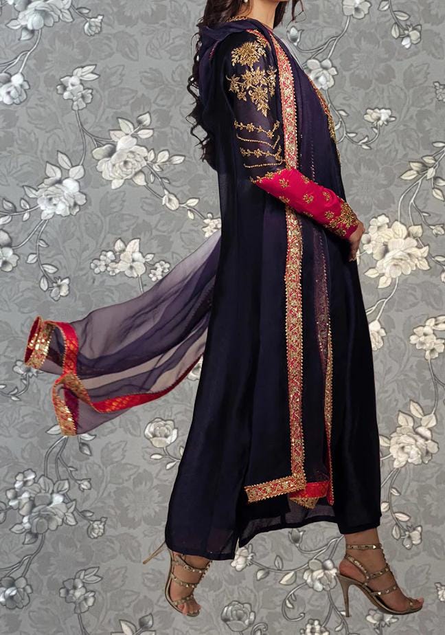 A woman wearing a long, dark blue dress with gold embroidery and red sleeves, standing against a gray floral background.