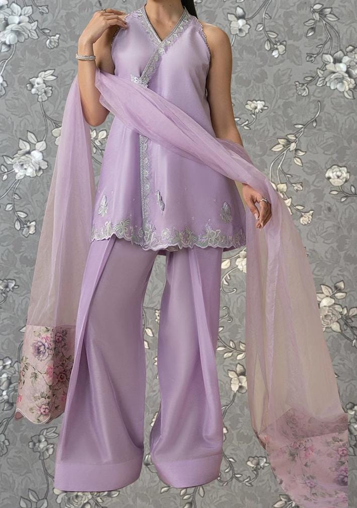 A woman wearing a lavender dress with a matching shawl, standing against a gray floral background.