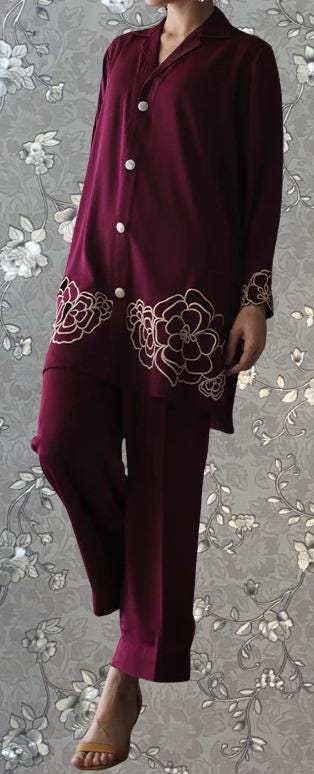 A person wearing a burgundy outfit with floral embroidery on the sleeves and pants, standing against a gray floral background.