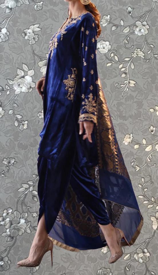 A woman wearing a long, blue dress with gold embroidery and a matching gold shawl, standing against a gray floral background.