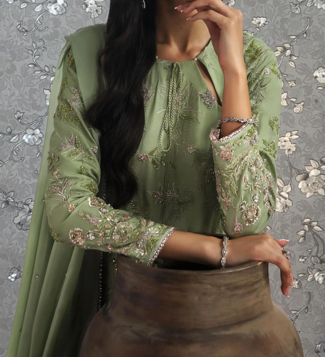 A woman wearing a green dress with intricate embroidery poses against a gray floral background.