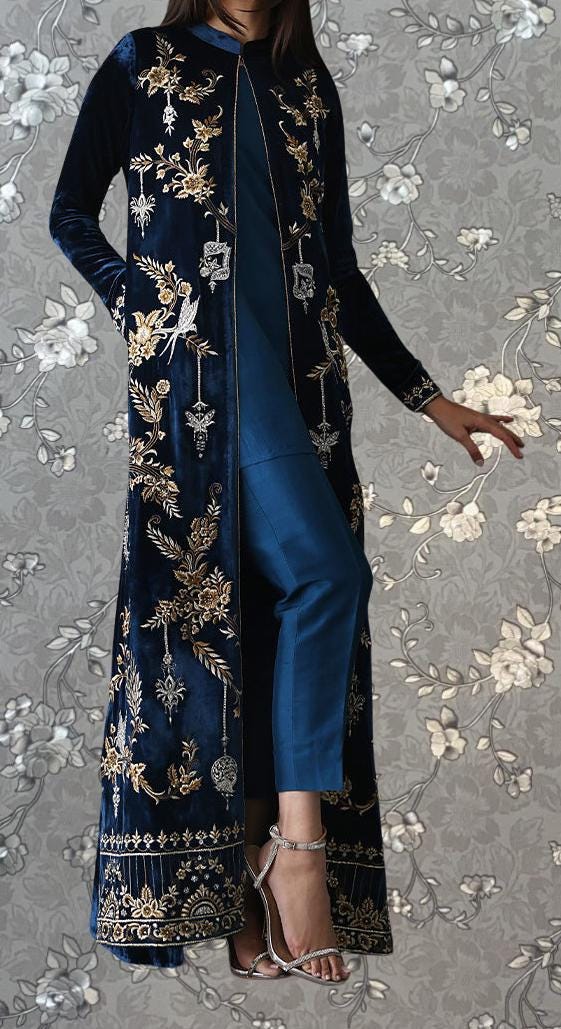 A woman wearing a long, blue velvet coat with gold embroidery stands against a gray floral background.