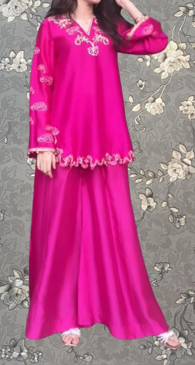 A woman wearing a long pink dress with intricate embroidery stands against a floral background.