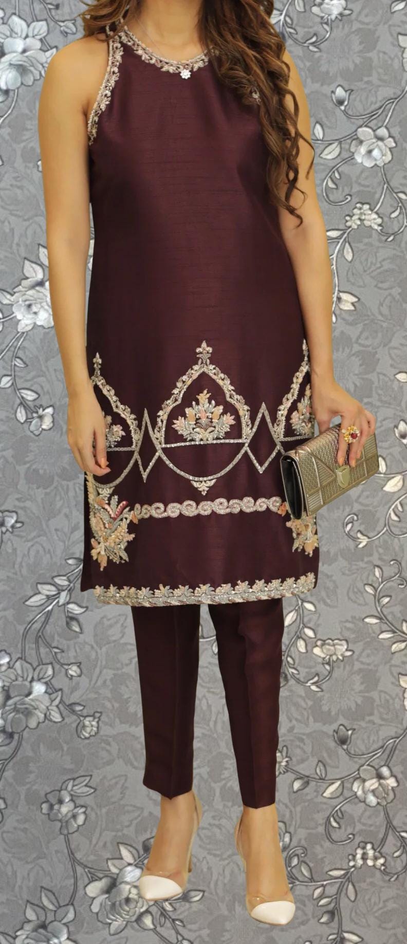A woman wearing a burgundy dress with intricate embroidery and holding a small clutch purse stands against a gray floral background.