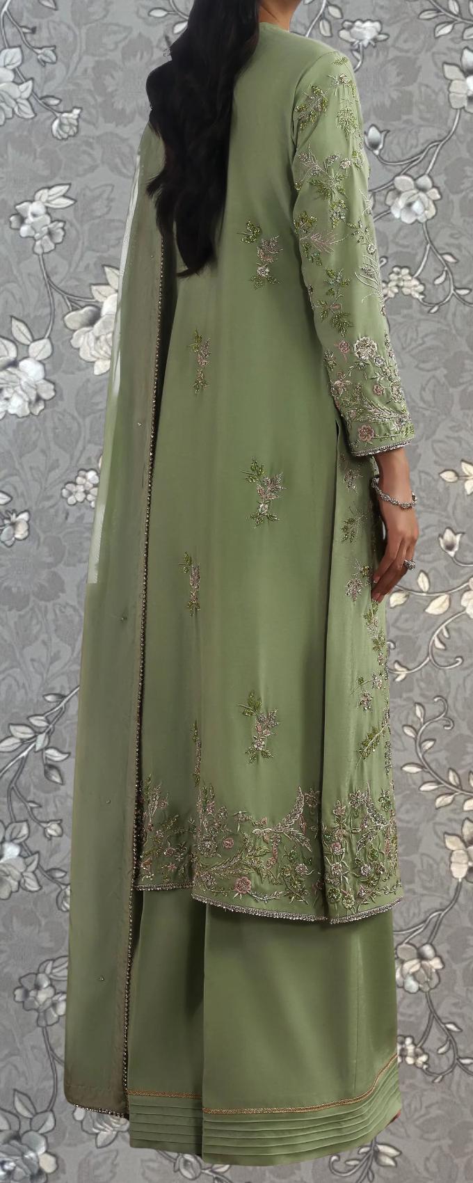 A woman wearing a green dress with intricate embroidery on the sleeves and skirt, standing against a gray floral background.