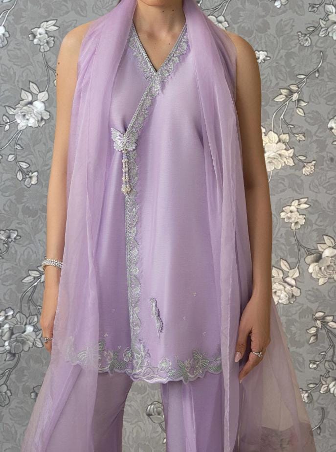 A woman wearing a purple dress with a sheer overlay and a matching scarf stands against a gray floral background.
