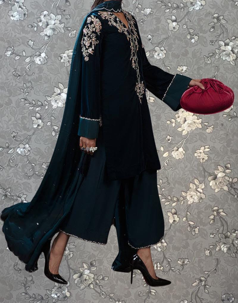 A person wearing a long black dress with intricate embroidery and a matching black purse is standing against a gray floral background.