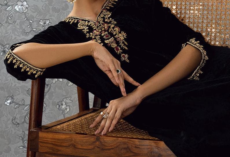 A woman wearing a black dress with gold embroidery and jewelry is sitting on a wooden chair, with her hands resting on the armrest.