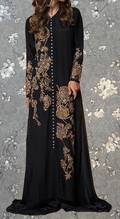 A woman wearing a long black dress with gold embroidery and buttons, standing against a gray floral background.