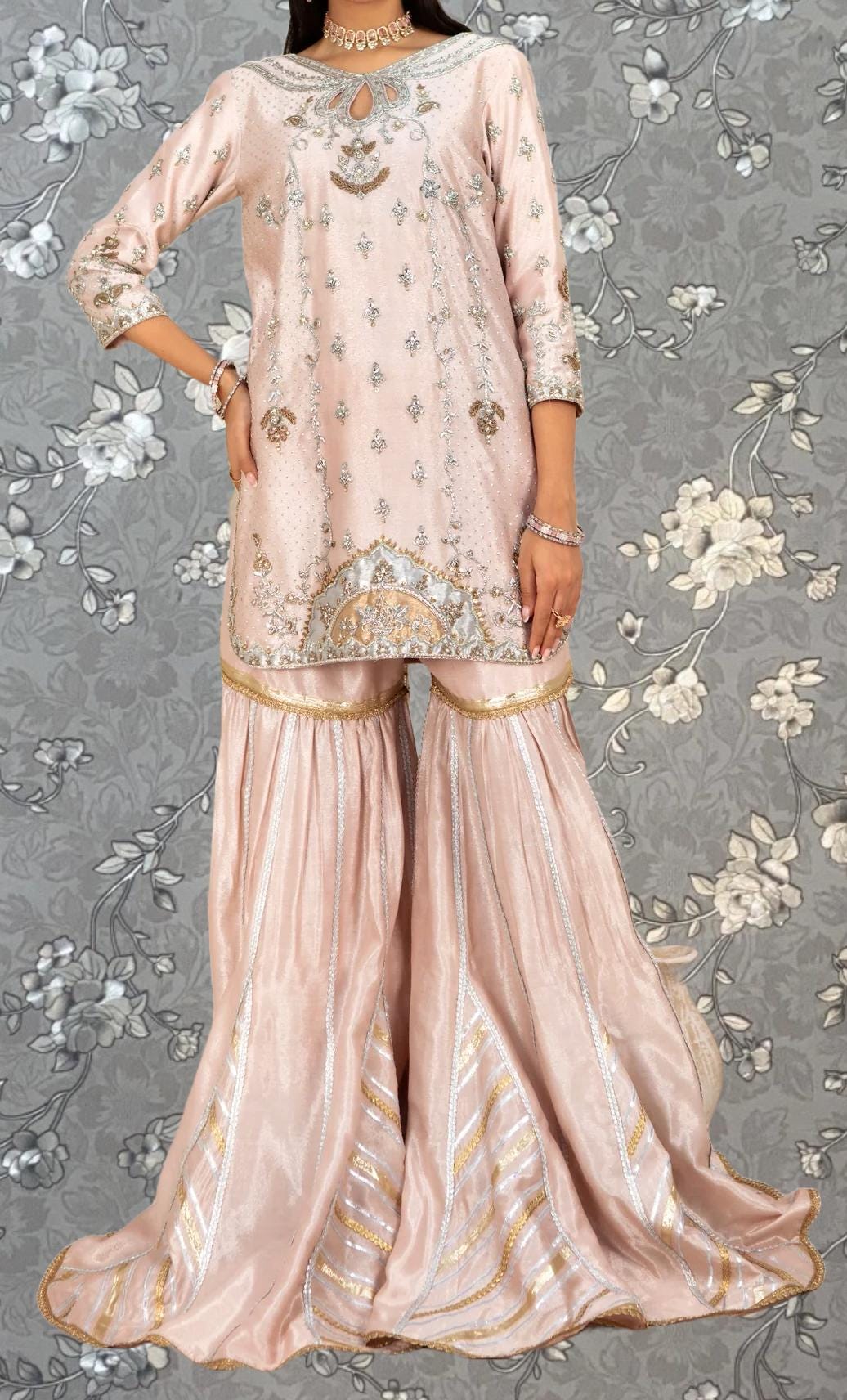 A woman wearing a pink and gold dress with intricate embroidery and a matching skirt, standing against a gray floral background.