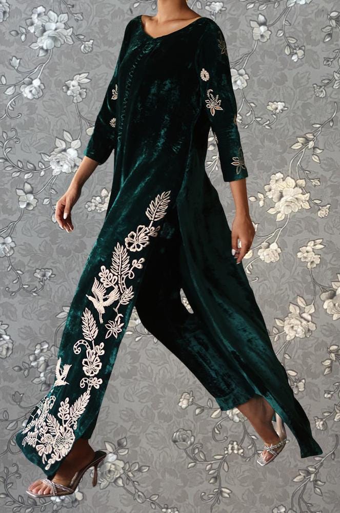 A person wearing a long, green velvet dress with white floral embroidery on the sleeves and legs, standing against a gray floral background.